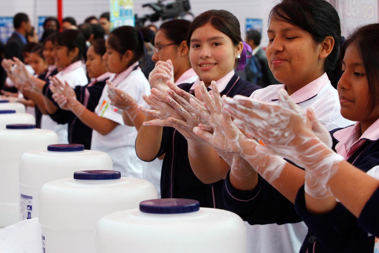  Image shows a line of girls in school uniforms rubbing their hands to a foamy lather with soap. 