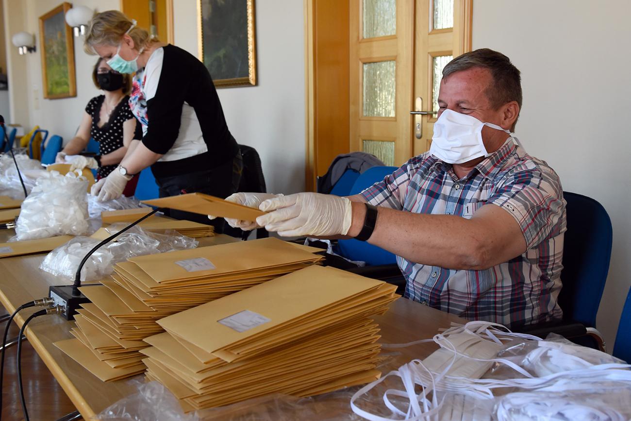 The photo shows a few city employees sitting at a table stuffing stacks of envelopes with masks. 
