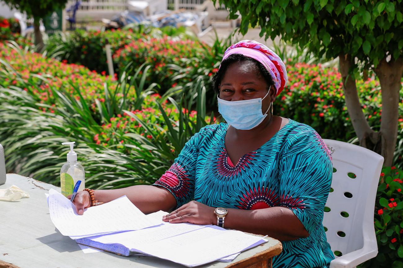 The photo shows a woman seated with a pad of paper on a sunny day in front of a garden full of flowers.