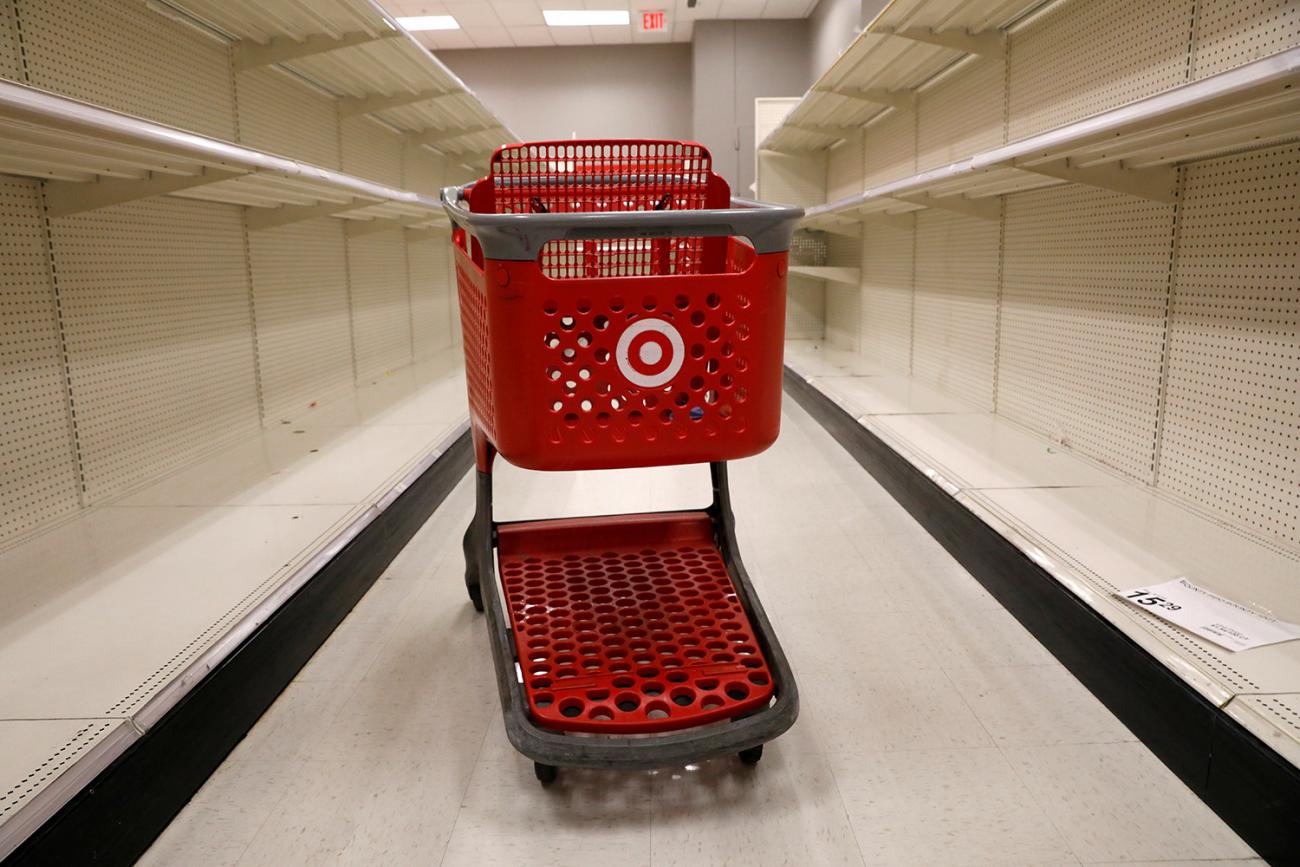 The photo shows a red shopping cart in between a broad aisle of empty shelves. 
