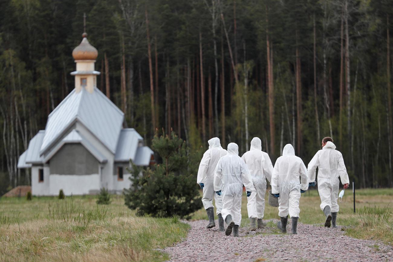 The photo shows a group of grave diggers wearing personal protective equipment walking weith their backs to the camera towards an old church. 