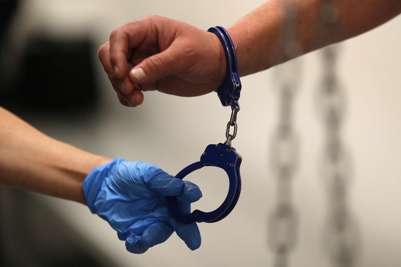 The photo shows a hand with a glove reaching for the uncuffed end of a pair of handcuffs attached to a hand with no glove. 