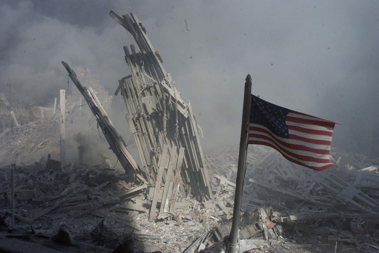 The photo shows a piece of the tower base shorn off at ground level with the American flag waving through the grim haze in the foreground. 