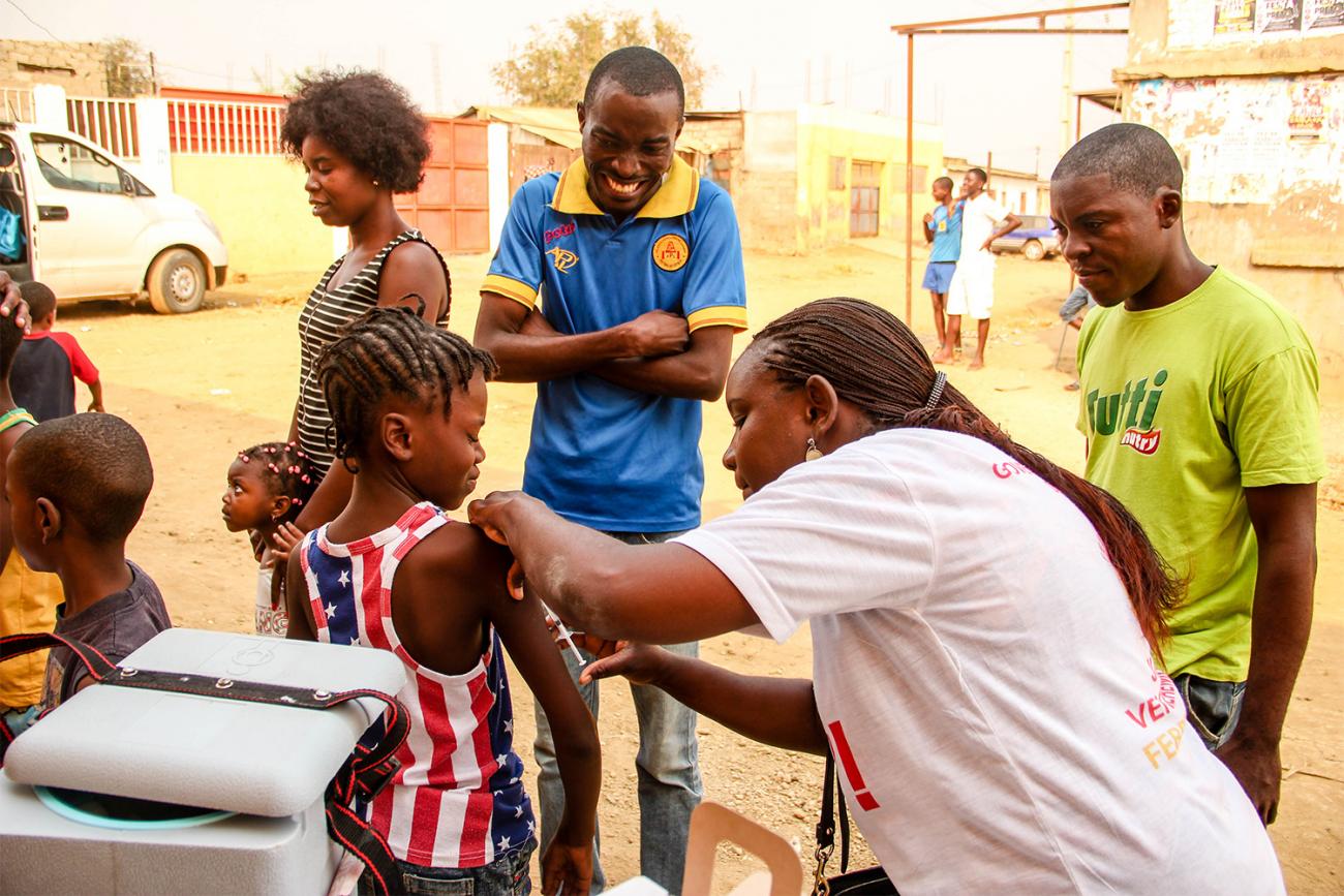 The photo shows a health worker administering a vaccine to a young girl as others look on.