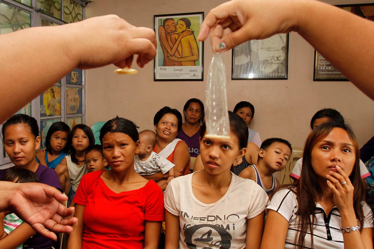 Picture shows the hands of a health worker holding up two condoms—one rolled, one unrolled—in front of a large audience of yound Philipino women. 