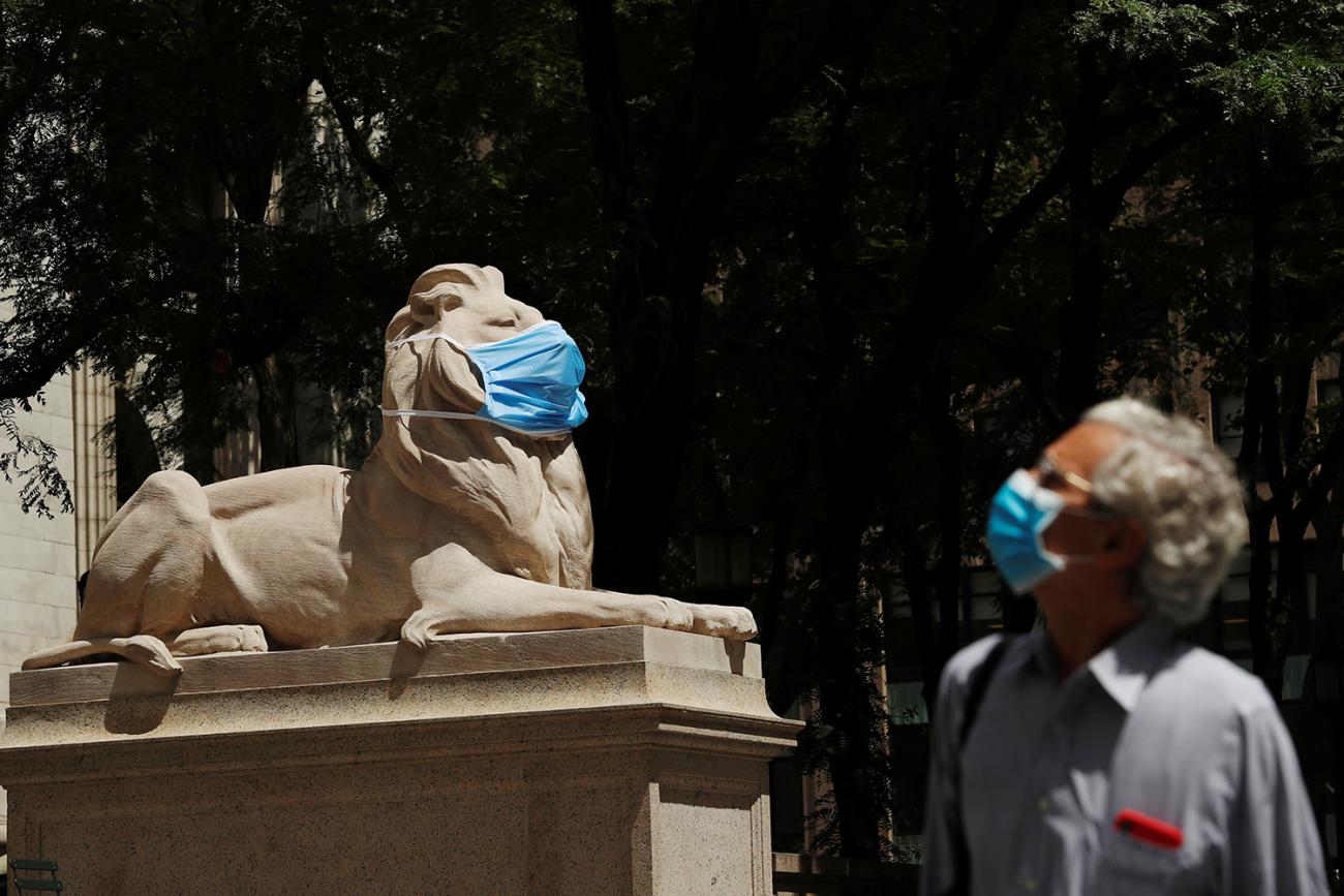 The photo shows a man in a blue face mask looking up at a stone sculpture of a lion also wearing a mask. 
