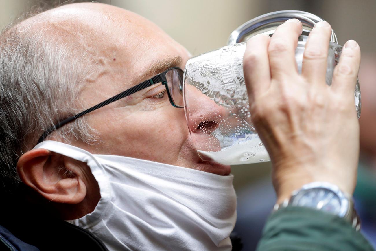 The photo shows a man with a mask pulled down around his chin gulping from a glass mug filled with frothy yellow liquid. 