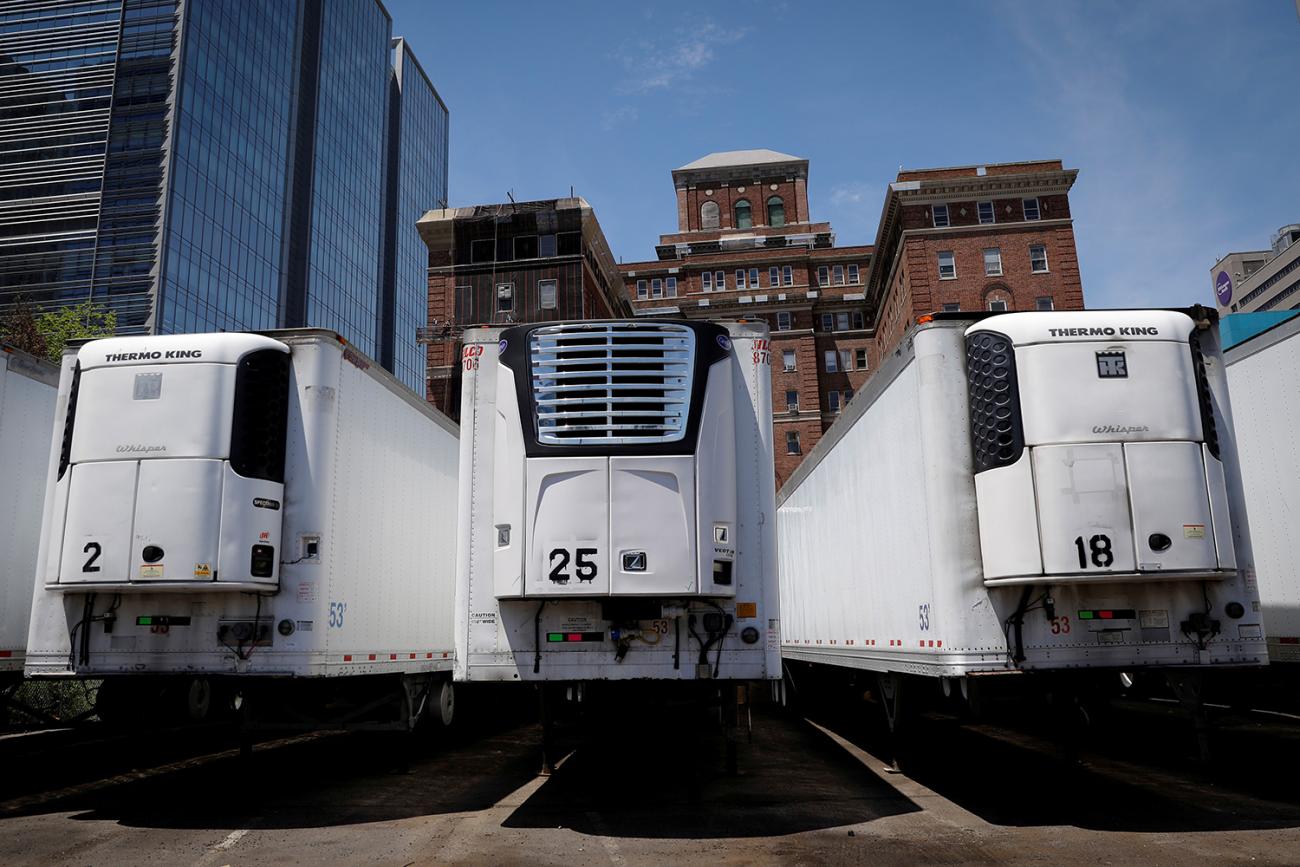 Image shows a row of huge refrigerated trucks parked together amid buildings under a clear blue sky. 