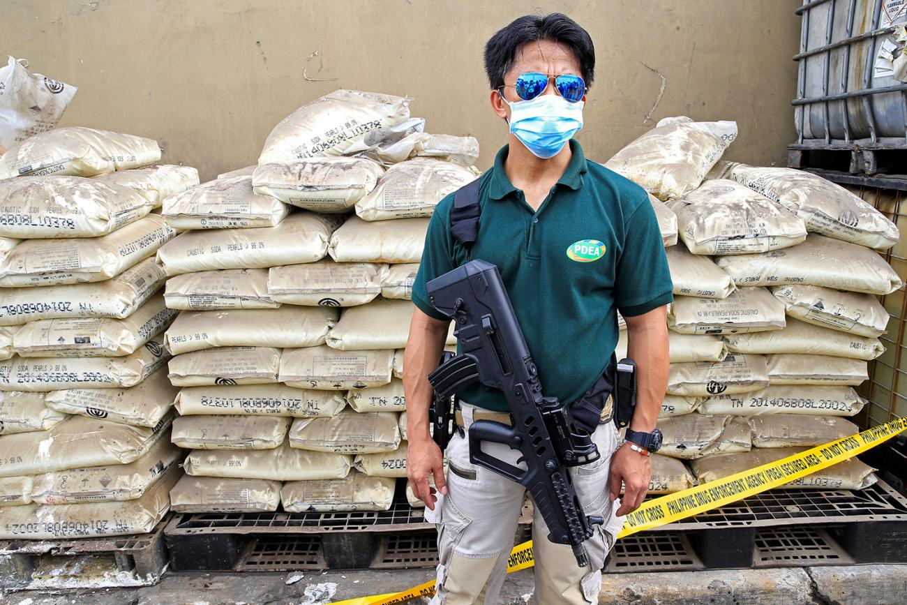 The photo shows the agent in reflective glasses standing in front of a huge stack of bagged chemicals. 
