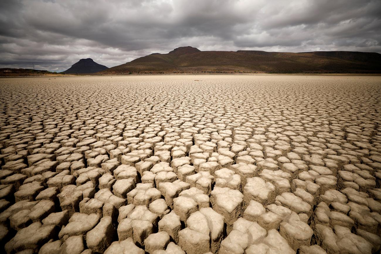 The dramatic photo shows a large mud plain dried to a cracked finish by drought and sun under a grey cloudy sky.