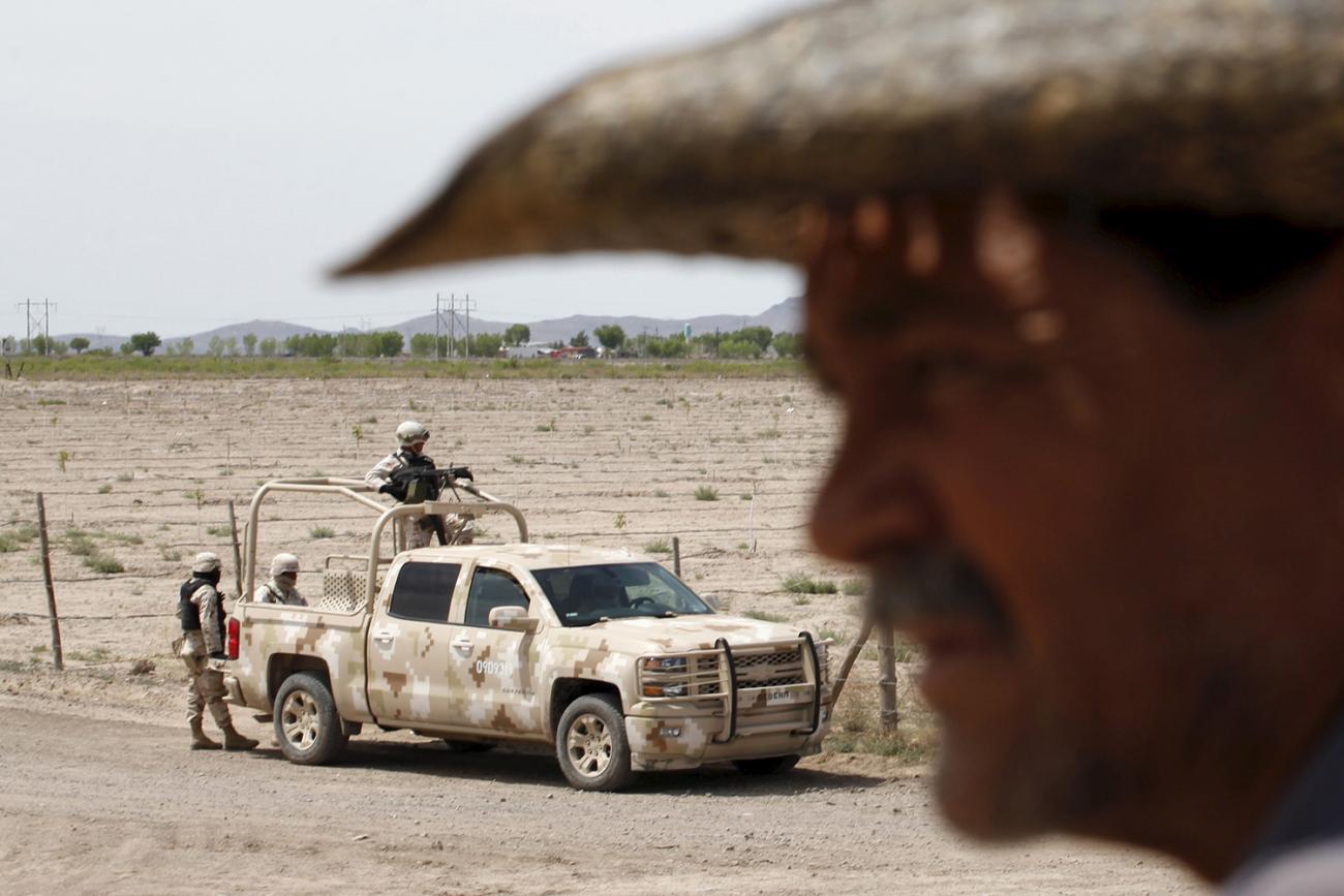 The photo shows a dusty dirt road with a truck of soldiers on it while a man stands out of focus in the foreground. 