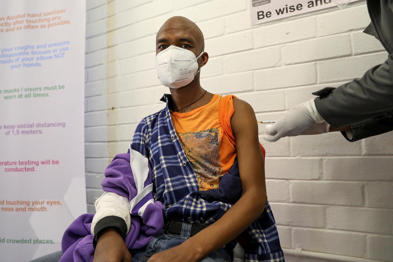 The photo shows a man wearing a mask with his sleeve rolled up taking an injection in the upper arm. 