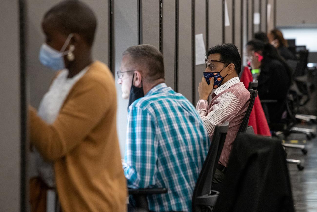 The photo shows an office with a row of cubicles and people actively making calls inside them. 