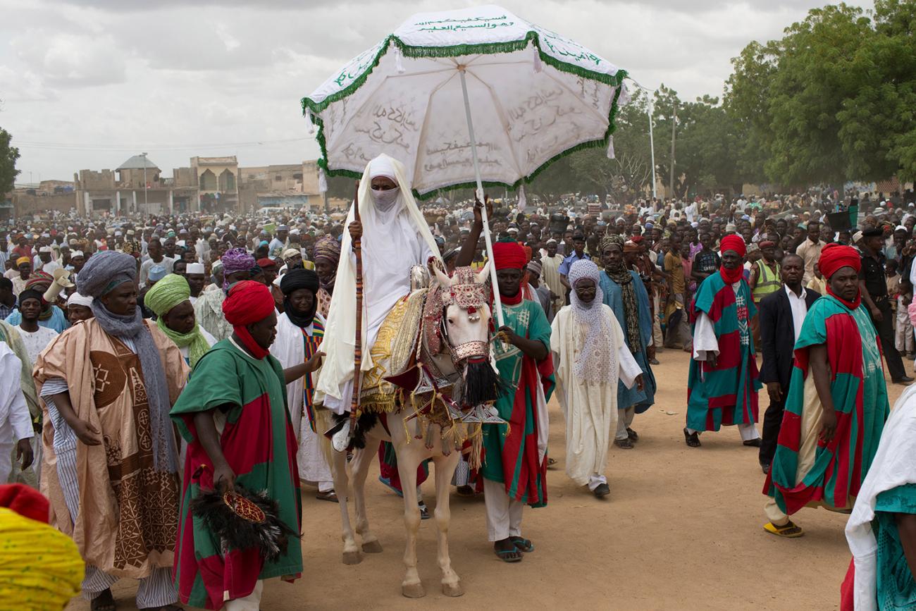 The photo shows the emir atop a horse with decorative coverings while a massive crowd surrounds him and one attendant holds an umbrella high above the emir's head. 