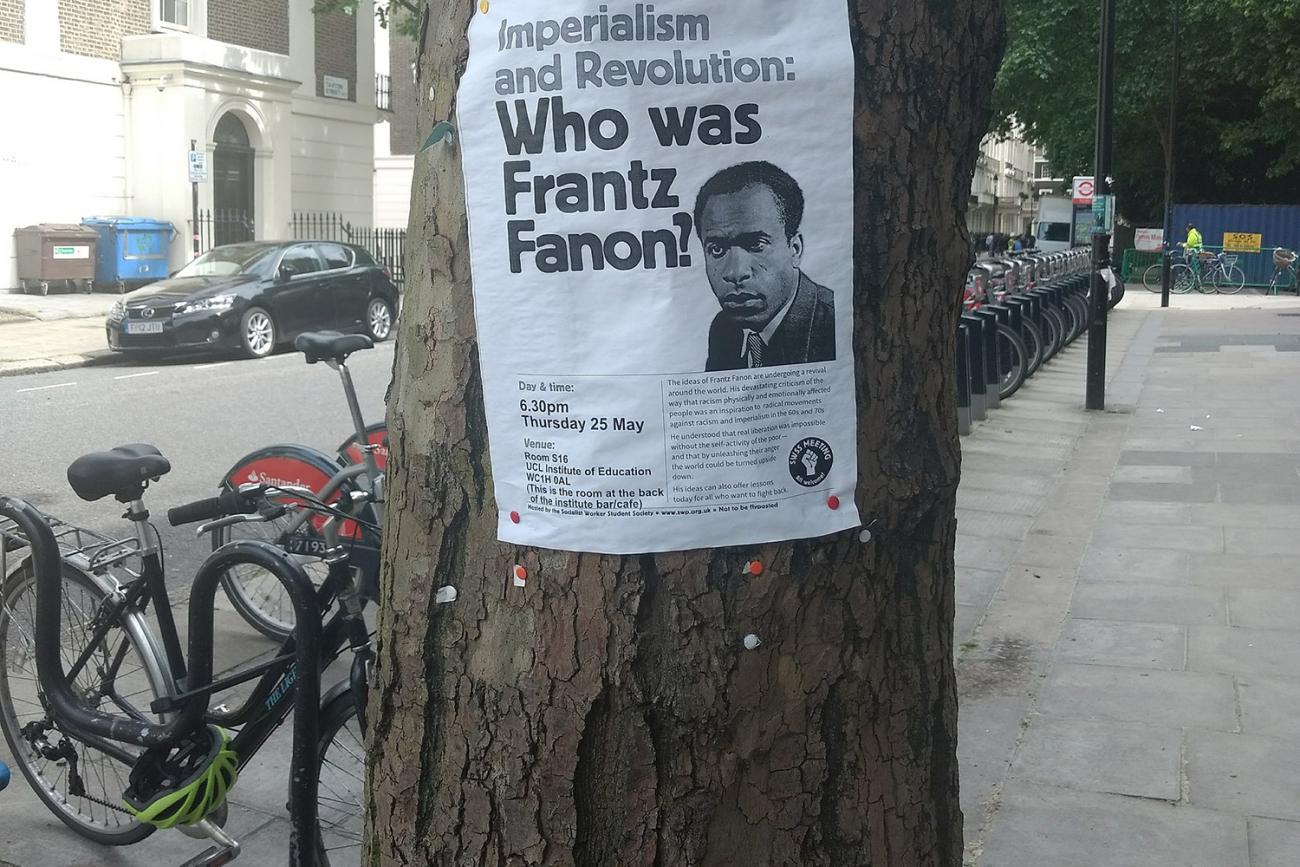 The photo shows a paper flier pasted to a tree that reads, "Who was Frantz Fanon?"