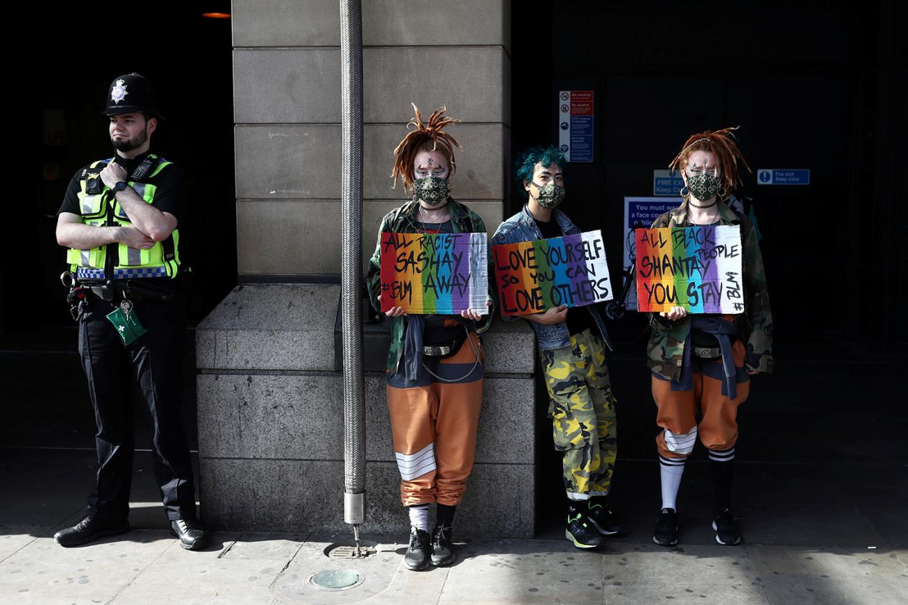 The photo shows three people in very fashionable protest wear lined up against a wall holding hand-made posters with a uniformed police officer next to them. 