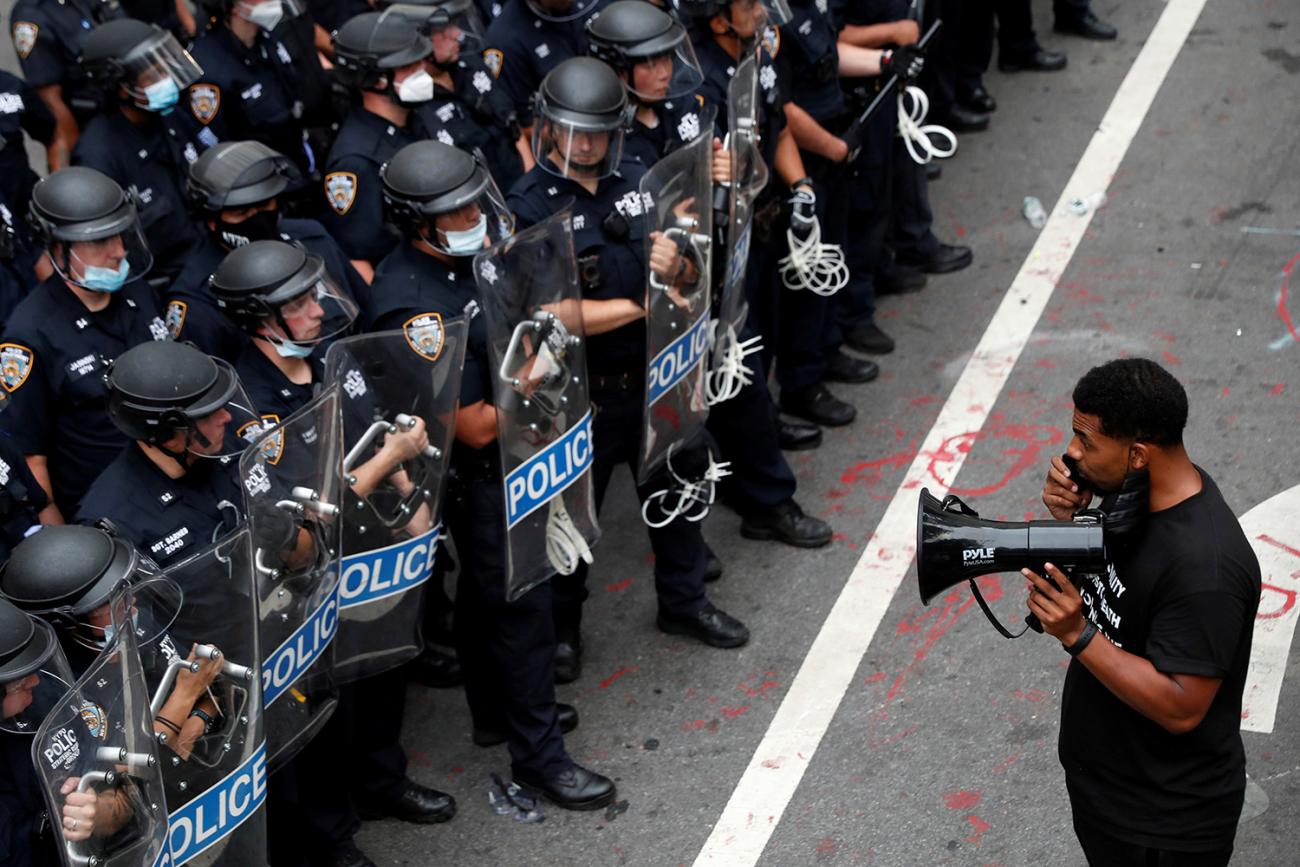 The photo shows a long, thick line of officers with one single protester holding a bull horn facing them and talking into the mic. 