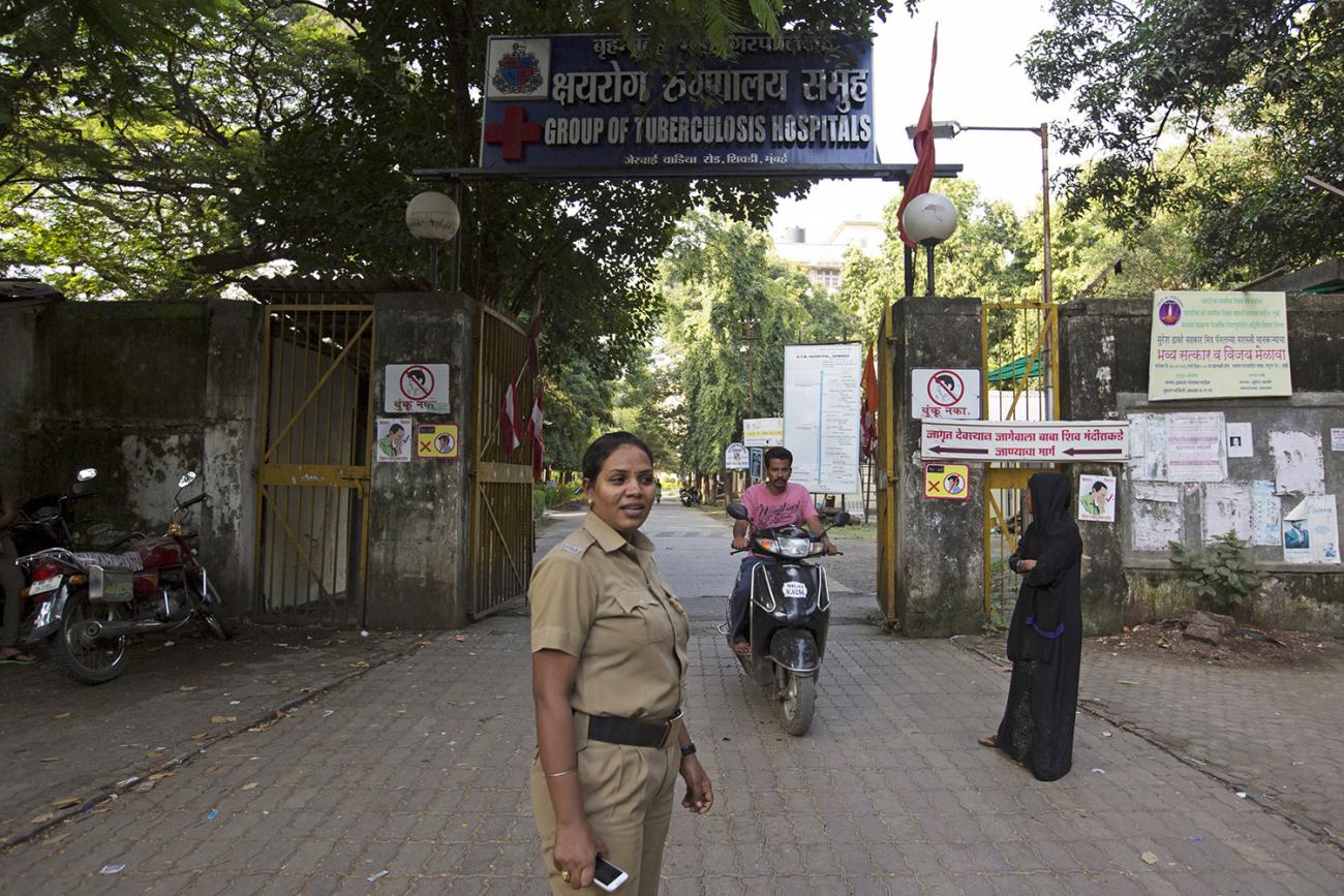 The photo shows a woman in a uniform by the gates of a hospital. 