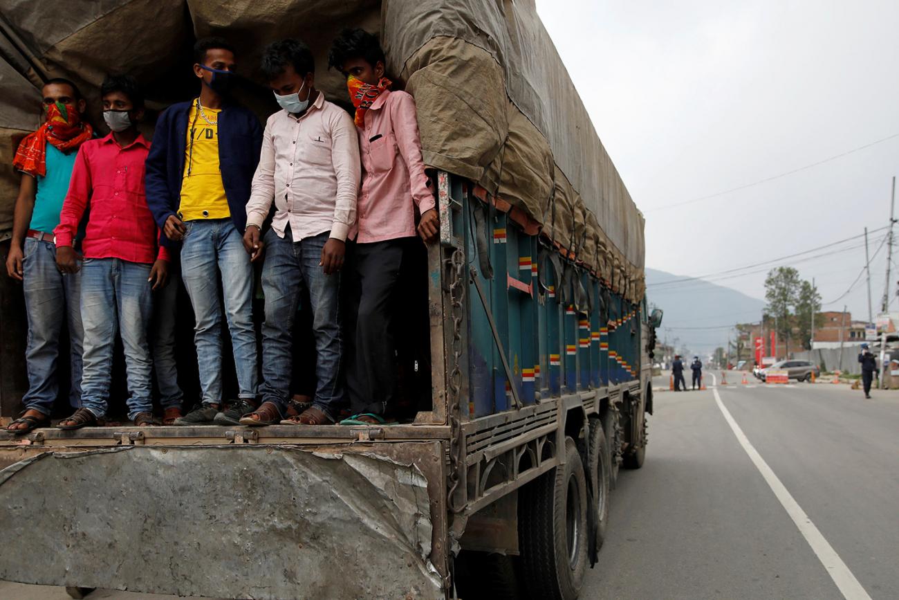 The photo shows a large flatbed trick covered with sides and a green tarp on a road. standing in the back of the truck can be seen several men. 