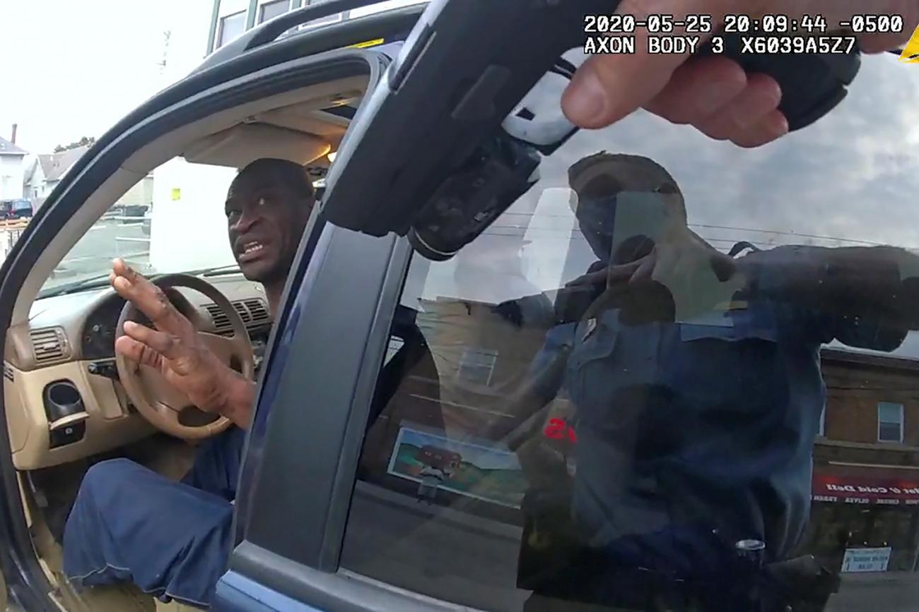 Image shows a single frame of a video with the first person perspective of an officer approaching Floyd, weapon visible in the foreground, as Floyd looks out from the front seat of his car. 
