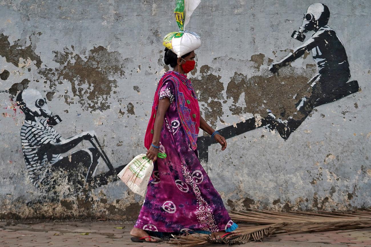 The photo shows a woman carrying a bag with another bag filled with what appears to be salt rested on her head. in the background is a mural of two children on a see-saw wearing full-on WWI-style gas masks.