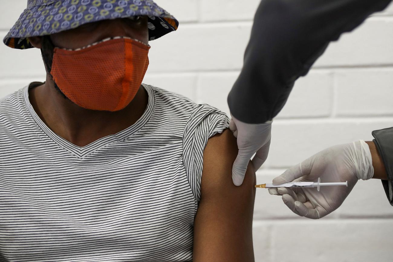 The photo shows a man with a hat and a mask getting a jab in his arm. 
