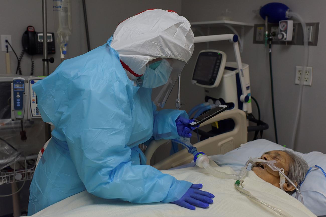 Picture shows a woman dressed in protective gear leaning over an older woman in a hospital bed with her phone in hand.