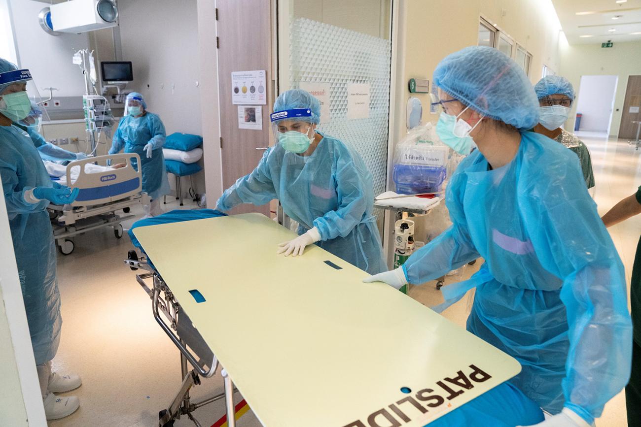 Picture shows sevreral health workers wearing blue protective equipment pushing a hospital gurney. 