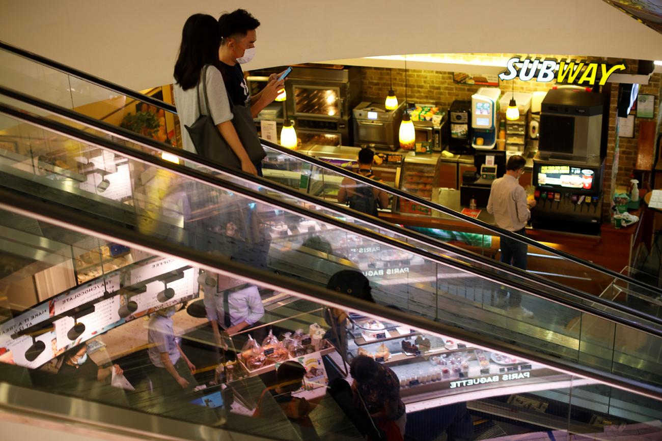 The photo shows a multi-level mall with several young people riding an escalator in the foreground. 