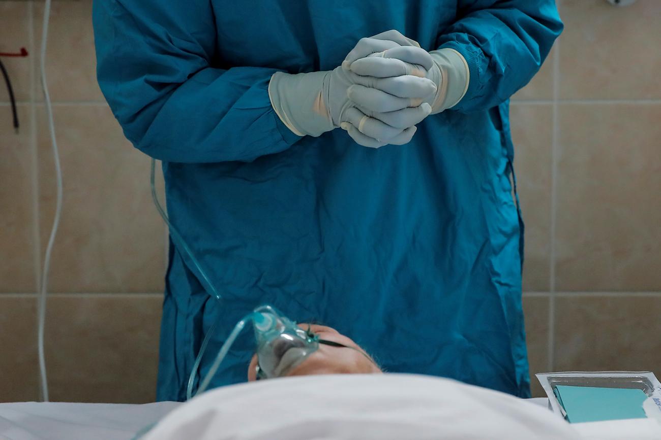 Picture shows a patient lying on their back on a hospital bed while the torso of a worker wearing protective gear and gripping hands together tightly can be seen standing over the patient. 