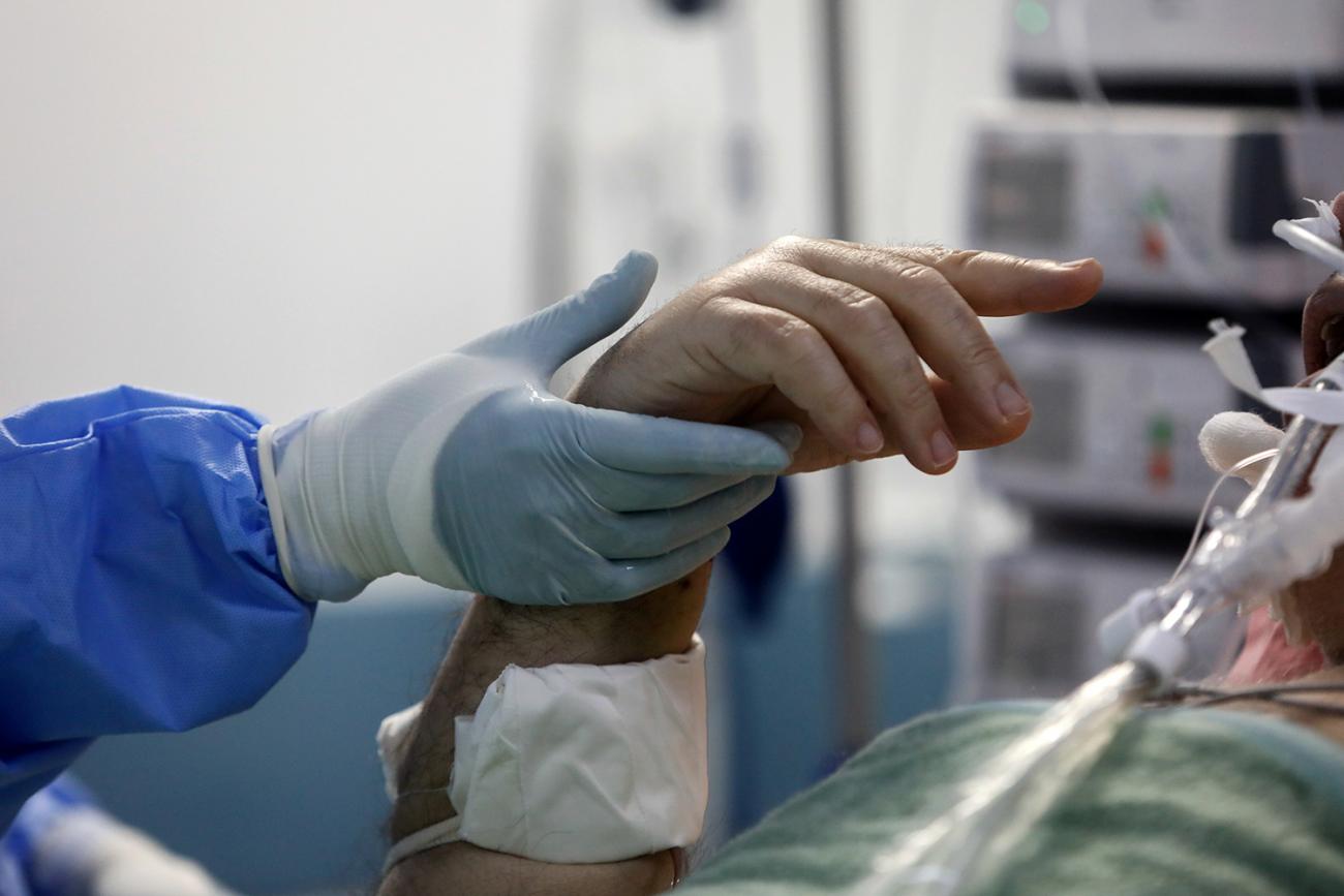 Picture shows the hand of a patient in a hospital bed being held by the blue-gloved hand of a medical worker. 