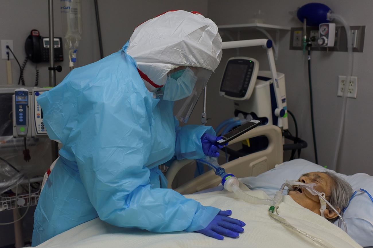 Picture shows a woman dressed in protective gear leaning over an older woman in a hospital bed with her phone in hand. 