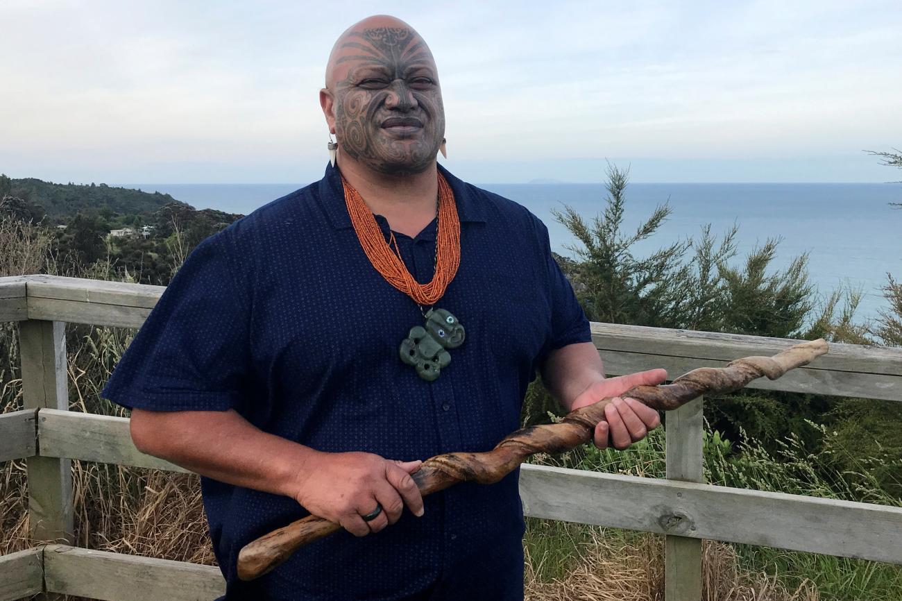 A Māori man with traditional face-tattoos stands in front of a wooden railing wearing a dark blue t-shirt