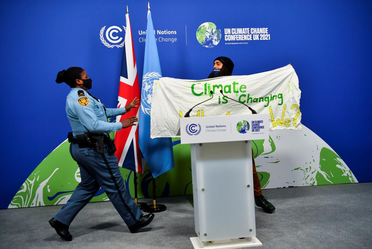 A security guard ushers a protester offstage. The protester is holding a banner that reads, "Climate is changing. Why Aren't We?"