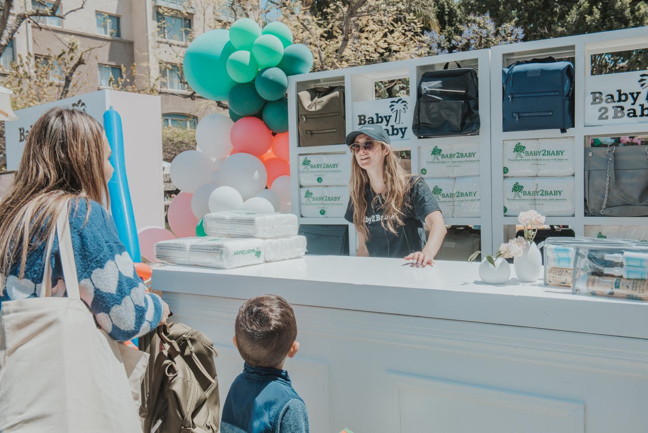A Baby2Baby community distribution event pictures an outdoor booth where a young woman volunteer behind a booth is greeting a mom and her little boy and girl, in Los Angeles, California, in 2022.
