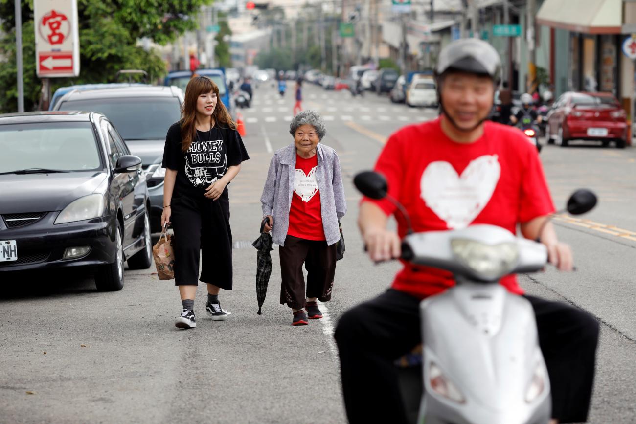 In the background, Wang-Chen Bi-Jin, 91 years old, a member of the "Six Carat" elderly Hip Hop dance group, walks with her coach Liu-Xin-Ru after a practice session. In the foreground, another member of the group rides a silver scooter. Both members are wearing red t-shirts with a white hearts in Taichung, Taiwan April 24, 2018. 