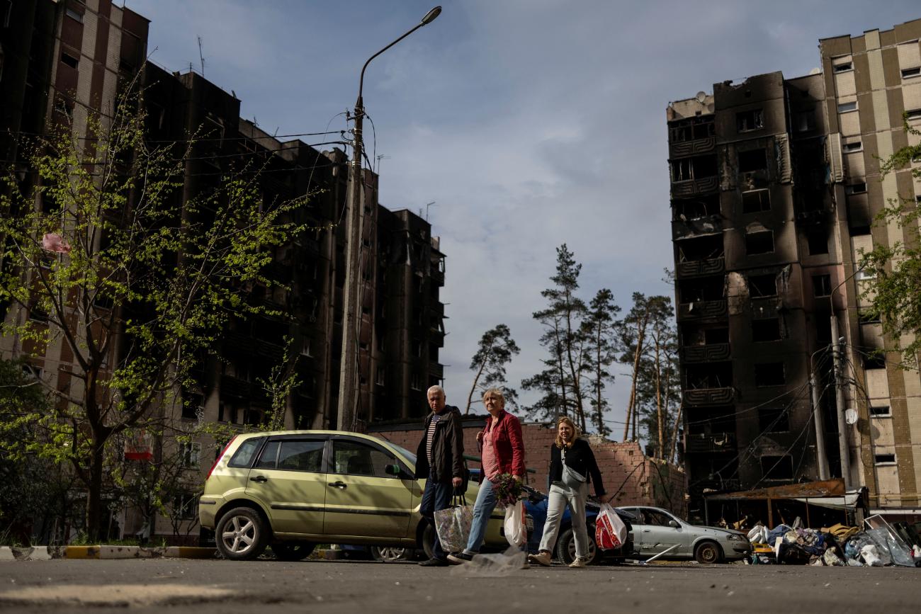 Several local residents carrying shopping bags walk by a small yellow car and in the background stand burned out buildings damaged by Russian shelling in a residential area of Irpin, Ukraine, on May 7, 2022.