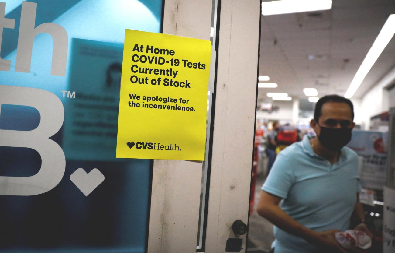 A man in a turquoise polo and mask passes the turquoise door of a CVS pharmacy, upon which is a yellow poster that reads, "At home COVID-19 tests currently out of stock. We apologize for the inconvenience."