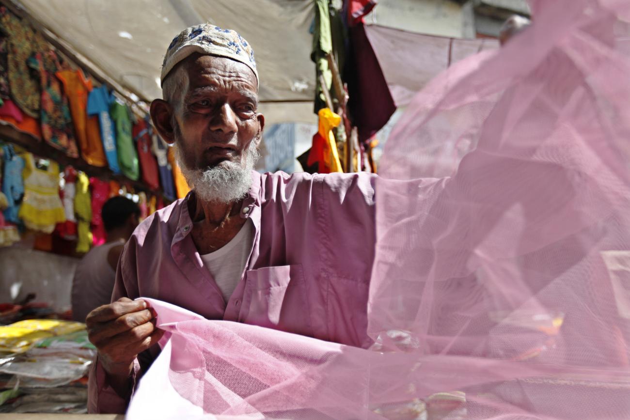 A tailor with a tan, aged face and short white beard wearing a pink shirt holds up a pink mosquito net at a market stall. His expression looks mildly concerned.