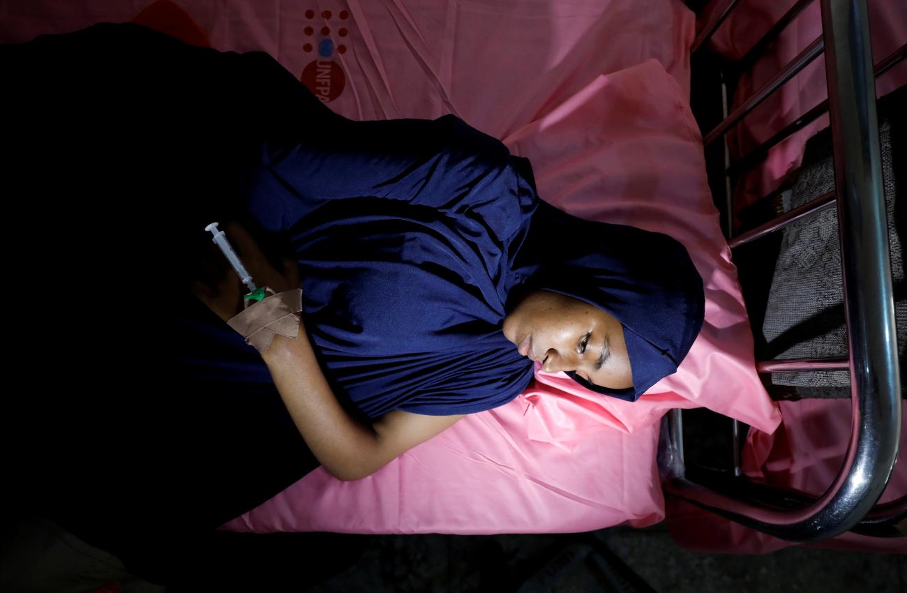 A young Nigerian woman in a navy dress and head scarf lies on a bed with pink sheets looking pensive as she recovers from surgery