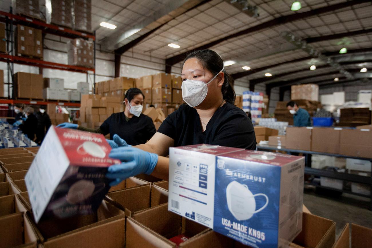 A woman in a black polo and white mask packs red and blue boxes of masks into larger brown cardboard boxes at a warehouse. In the background, other workers do the same.