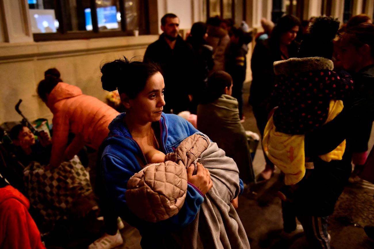 A mother in a blue jacket with a worried expression on her face breastfeeds her baby on a busy train platform at night. In the background other refugees bustle with luggage and children.  