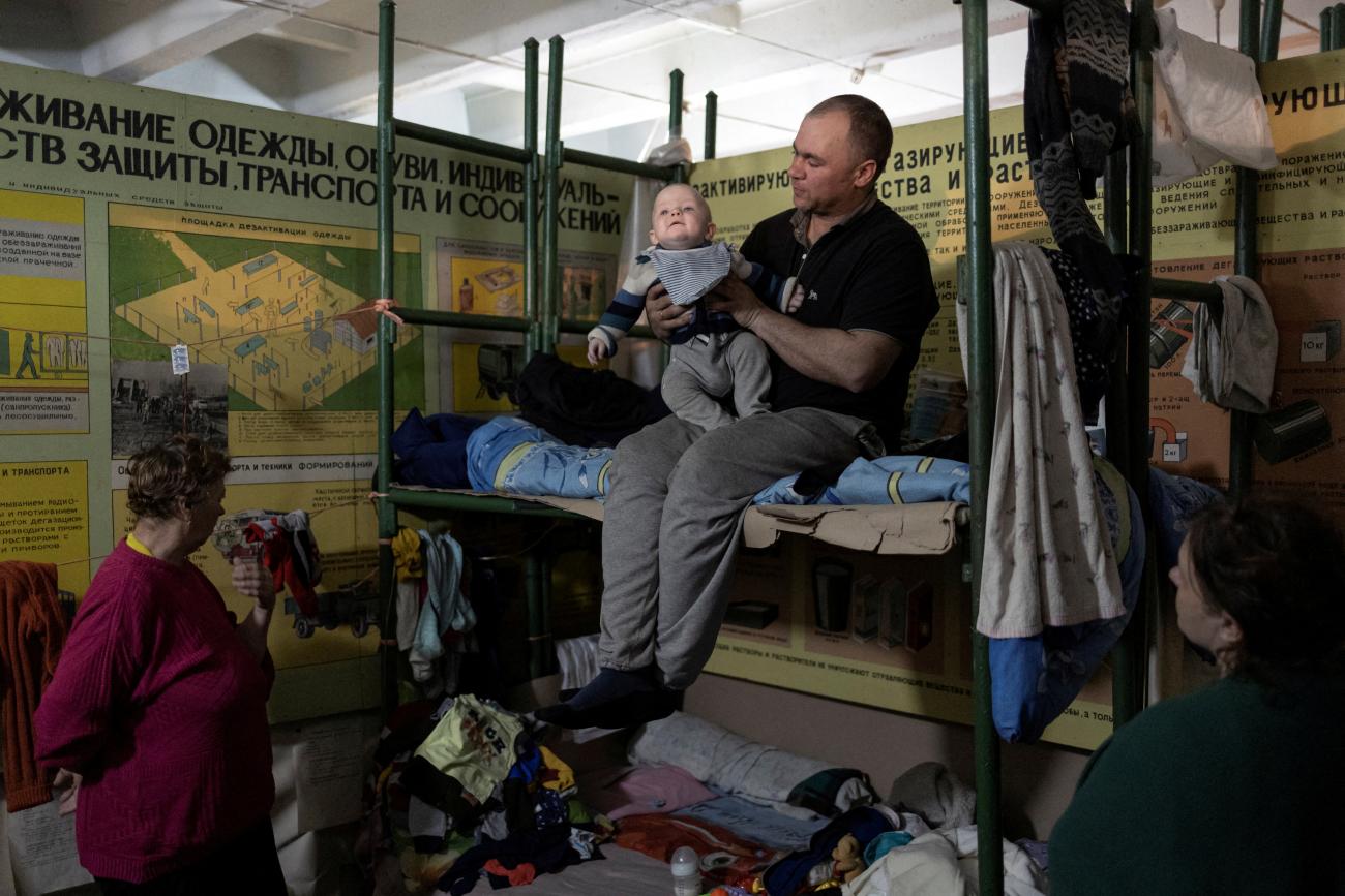 A balding man sits on a makeshift bunk bed holding a baby in a dimly lit bomb shelter, surrounded by clothes and blankets of many colors. In the foreground, two women stand in shadow, looking at the man and baby. 