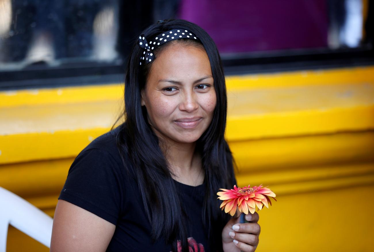 A woman in a black t-shirt with long dark hair stands against a yellow background and holds a pink and orange flower as she waits to be released from jail after the Supreme Court of El Salvador commuted her sentence for abortion, in IIopango, El Salvador on March 7, 2019. 