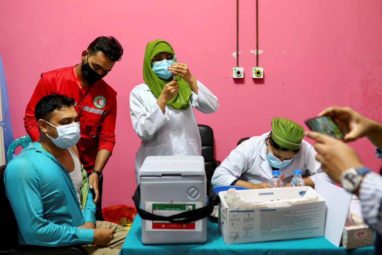Female doctors in white lab coats and green head scarves prepare a vaccine for a young man in a turquoise sweatshirt in a room with pink walls