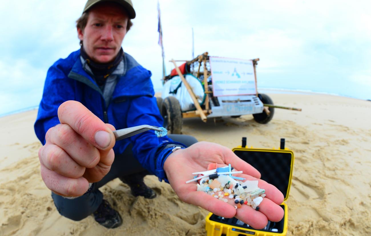 French scientist Edgar Dusacre, wearing a baseball cap and blue windbreaker, squats on a beach holding his palm out, which is full of microplastic waste collected on the Aquitaine coast on the beach of Contis, in southwestern France, on August 17, 2020.