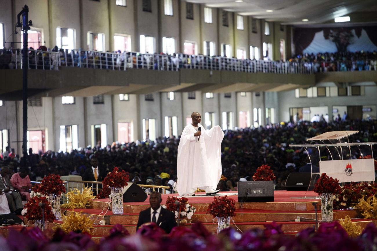 Bishop David Oyedepo wearing white religious garments leads a sermon in a full auditorium at a church