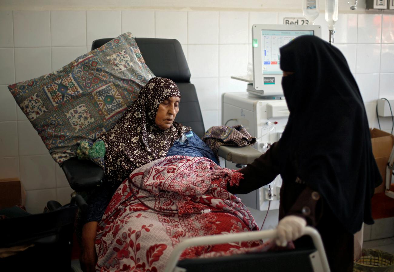 A woman in a floral head scarf sits propped up on patterned pillows and covered with ornate blankets, looking tired as she undergoes dialysis treatment. To her right a woman in all black holds her hand