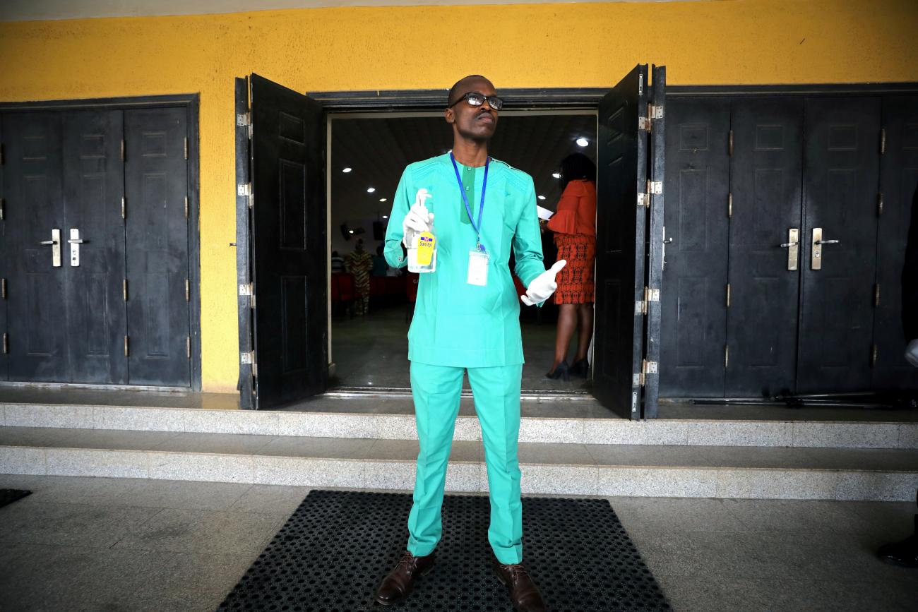 A man wearing bright blue traditional clothing stands in front of a church entrance holding a canister of hand sanitizer