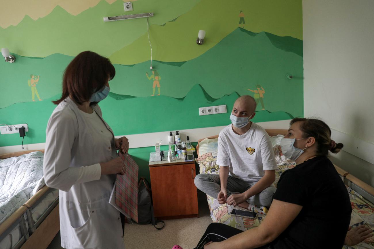 A young cancer patient and her mother sit on a bed facing an oncologist in a room with a green mural of mountain climers scaling a mountain. 